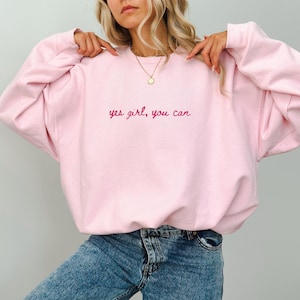 Könnte beinhalten: Ein hellrosa Sweatshirt mit langen Ärmeln. Der Text "yes girl, you can" ist in einer Schreibschrift auf die Brust gestickt. Das Sweatshirt wird mit einer blauen Jeans kombiniert.