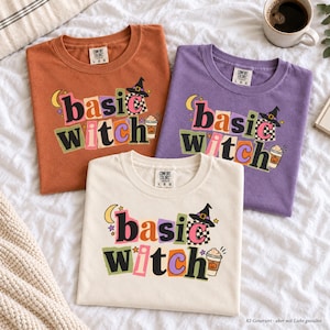 Könnte beinhalten: Drei Rundhals-T-Shirts in Rostorange, Lila und Creme, jeweils mit dem Schriftzug "basic witch" in bunten, blockartigen Buchstaben. Die Grafik zeigt einen schwarzen Hexenhut, einen gelben Halbmond, kleine Sterne und einen Kaffeebecher mit Kürbis.