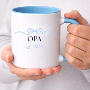 May include: White ceramic mug with a light blue interior and handle. The mug features the word "OPA" in navy blue, with "est. 2026" below it, and a light blue bow design above. The mug is held by a person.