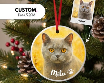 May include: A round ceramic Christmas ornament featuring a gray cat with yellow eyes, a red ribbon, and the name "Mila". The ornament has a watercolor-style image of the cat on a yellow background. A small photo of the cat is also included.