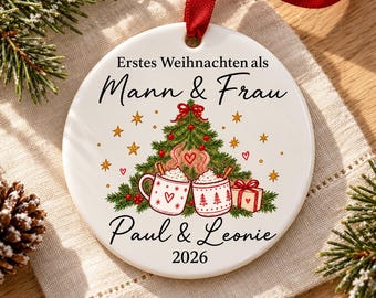 May include: A round white ceramic ornament with a red ribbon, featuring a printed Christmas tree, two mugs of hot cocoa, and a gift box. The text reads: Erstes Weihnachten als Mann & Frau, Paul & Leonie, and the year 2026 in black lettering.