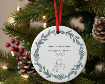 May include: A white ceramic Christmas ornament with a red ribbon, hanging on a Christmas tree. The ornament has a green floral wreath design around the edge. Text on the ornament reads "Erstes Weihnachten im eigenen Zuhause 2025" with a key graphic.
