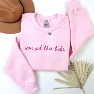 May include: A light pink sweatshirt with the phrase "you got this babe" in a darker pink script. A gold necklace with a small pendant is draped on the sweatshirt. A brown hat and dried palm leaves are in the background.