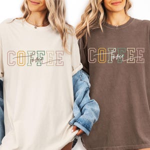 May include: Two t-shirts with the text "COFFEE lover" in a colorful, outlined font. One shirt is cream-colored, the other is brown. The word "lover" is written in a cursive font. The shirts are worn by a person.