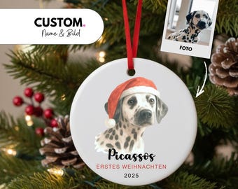 May include: A white ceramic Christmas ornament featuring a Dalmatian dog wearing a Santa hat. The ornament has a red ribbon for hanging and includes the text "Picassos, ERSTES WEIHNACHTEN 2025". A small photo of the dog is also included.