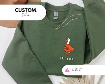 Könnte beinhalten: Ein olivgrünes Sweatshirt mit Rundhalsausschnitt und langen Ärmeln. Auf der Vorderseite ist eine Cartoon-Gans abgebildet, die einen orangefarbenen Pullover mit dem Text "Papa Goose" und "EST. 2025" trägt. Eine goldene Halskette ist über dem Sweatshirt drapiert.
