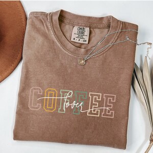 May include: A brown Comfort Colors t-shirt with the word "COFFEE" in a multi-colored block letter design and the word "lover" in a cursive font. A silver necklace with a small pendant is draped across the shirt.