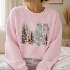 Könnte beinhalten: Ein hellrosa Sweatshirt mit Rundhalsausschnitt und vier vertikalen Aquarell-Paneelen. Die Paneele zeigen einen beleuchteten Weihnachtsbaum, einen Nussknacker-Soldaten, eine tanzende Ballerina und ein tanzendes Paar in einem Ballsaal.