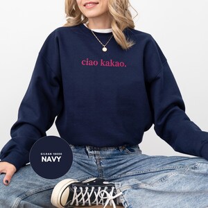 May include: Navy blue sweatshirt with the text "ciao kakao." in pink lettering. The sweatshirt is a crew neck style and is paired with blue jeans. The brand is Gildan 18000.