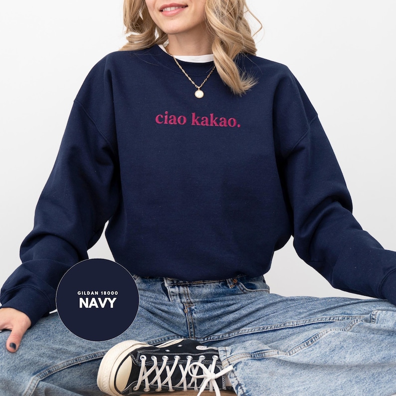 May include: Navy blue sweatshirt with the text "ciao kakao." in pink lettering. The sweatshirt is a crew neck style and is paired with blue jeans. The brand is Gildan 18000.
