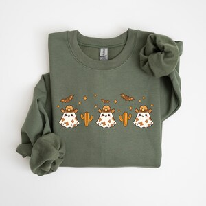 Könnte beinhalten: Ein olivgrünes Sweatshirt mit einem Design aus Cartoon-Geistern mit Cowboyhüten, Kakteen und Fledermäusen. Die Geister sind weiß mit braunen Flecken. Das Design enthält orangefarbene Sterne und Fledermäuse.