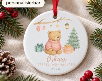 May include: A round white ceramic Christmas ornament with a red ribbon, featuring a watercolor illustration of a teddy bear in a pink sweater, a small Christmas tree, gifts, and hanging decorations, with the text Oskars ERSTES WEIHNACHTEN 2026.
