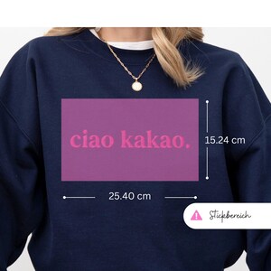 May include: Navy blue sweatshirt with a rectangular magenta design that reads "ciao kakao." The design measures 10 inches wide and 6 inches high. A gold necklace with a pendant is visible. The sweatshirt is a casual style.