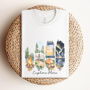 Könnte beinhalten: Ein weißes T-Shirt mit einer Aquarell-Grafik aus vier vertikalen Feldern, die ein Lagerfeuer, einen Wohnwagen, ein Zelt am See und Nordlichter zeigen. Der Schriftzug "Explore More" ist darunter in schwarzer Schreibschrift aufgedruckt.