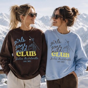 Könnte beinhalten: Ein dunkelbraunes und ein hellblaues Sweatshirt mit Rundhalsausschnitt, beide mit dem Text 'girls gone mild CLUB' in Blau und Gelb, darunter 'Julias Bachelorette' und 'LAAX, SWISS' sowie eine grafische Abbildung einer Skigondel.