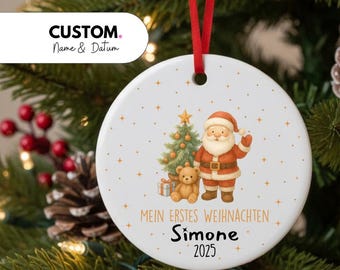 May include: A round white ceramic Christmas ornament hanging from a red ribbon. The ornament features a graphic of Santa Claus, a decorated Christmas tree, and a teddy bear, with the text "MEIN ERSTES WEIHNACHTEN", "Simone", and "2025".