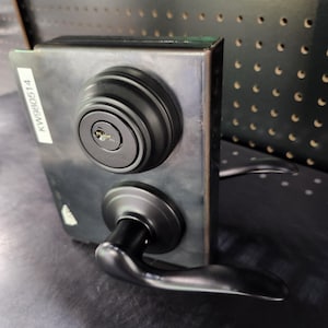 May include: A black door lock set with a keyhole and a handle. The lock is mounted on a dark metal plate. A white sticker with the text "KW980514" is on the left side of the plate. The handle is curved and positioned below the lock.