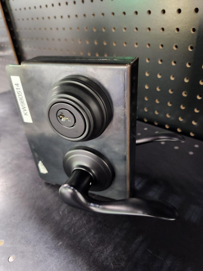 May include: A black door lock set with a keyhole and a handle. The lock is mounted on a dark metal plate. A white sticker with the text "KW980514" is on the left side of the plate. The handle is curved and positioned below the lock.