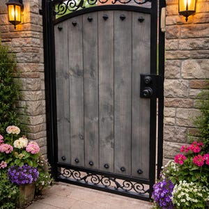 May include: A wooden gate with a gray finish, framed by black metal and adorned with decorative scrollwork. The gate is flanked by brick columns with lit lanterns and flower pots filled with pink, purple, and white flowers.