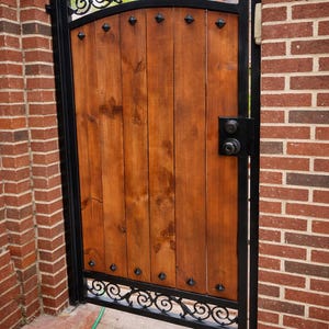 May include: A wooden gate with a black metal frame and decorative scrollwork. The gate is made of vertical wooden planks and features a black handle and hinges. The gate is set between two brick walls.
