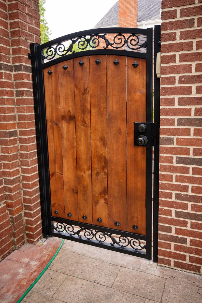 May include: A wooden gate with a black metal frame and decorative scrollwork. The gate is made of vertical wooden planks and features a black handle and hinges. The gate is set between two brick walls.