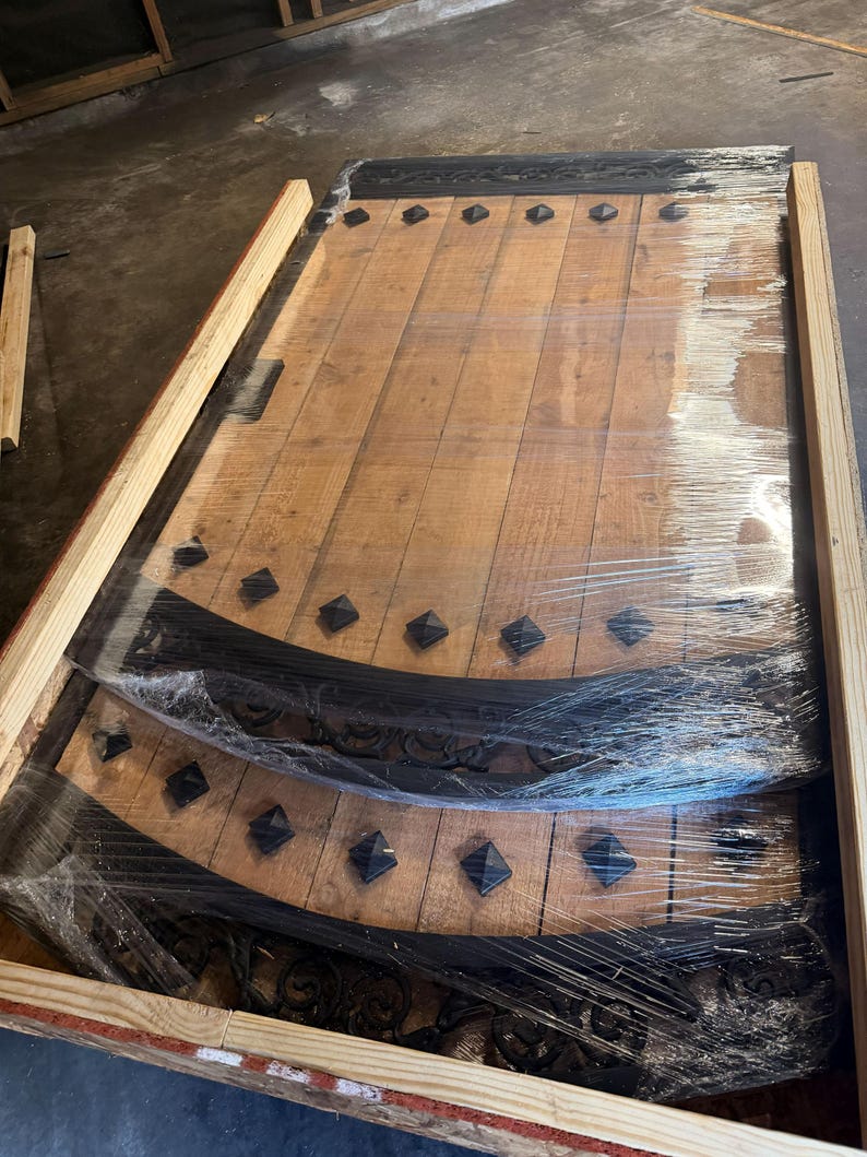 May include: Wooden door panels with decorative black metal accents and diamond-shaped studs. The panels are wrapped in clear plastic and are inside a wooden crate. The wood has a natural finish.