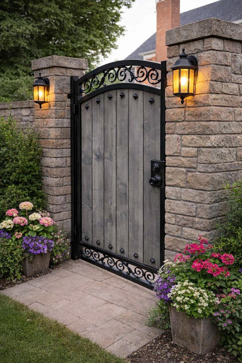 May include: A gray wooden gate with black wrought iron accents and a decorative top. The gate is flanked by stone pillars with sconce lights. Colorful flowers in stone planters line the walkway.