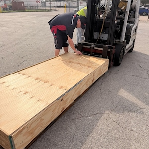 May include: A forklift lifting a large, rectangular wooden crate. Two people are attending to the crate. The crate is made of light-colored wood and has metal corner reinforcements. The scene takes place outdoors on a paved surface.