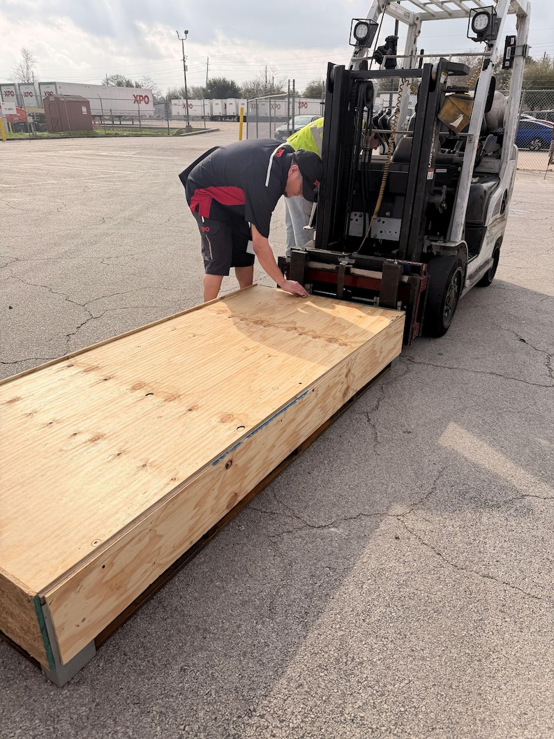 May include: A forklift lifting a large, rectangular wooden crate. Two people are attending to the crate. The crate is made of light-colored wood and has metal corner reinforcements. The scene takes place outdoors on a paved surface.