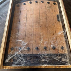 May include: A wooden door with a dark finish, featuring vertical planks and decorative black metal accents. The door has a curved top and is encased in a wooden crate, wrapped in clear plastic for protection. The door has black diamond-shaped studs.