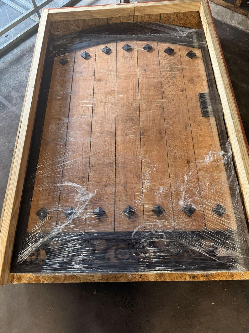 May include: A wooden door with a dark finish, featuring vertical planks and decorative black metal accents. The door has a curved top and is encased in a wooden crate, wrapped in clear plastic for protection. The door has black diamond-shaped studs.