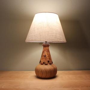 May include: A wooden table lamp with a beige linen lampshade. The lamp base is crafted from light-colored wood, featuring a bulbous bottom and a detailed, textured design on the upper portion. The lamp is on a wooden surface.