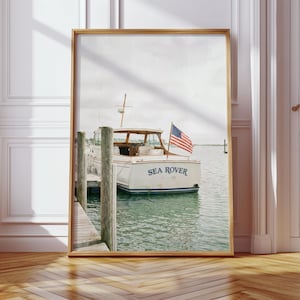 Peut inclure: Impression encadrée d'un bateau blanc nommé "Sea Rover" avec un drapeau américain, amarré à un ponton en bois. Le bateau est sur l'eau et le ciel est nuageux. L'impression est dans un cadre doré.
