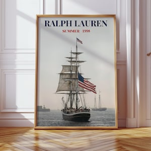 Puede incluir: Una impresión enmarcada que presenta un velero navegando sobre el agua, con la bandera estadounidense ondeando. La impresión tiene el texto "RALPH LAUREN SUMMER 1998" en la parte superior. La impresión está en un marco dorado.