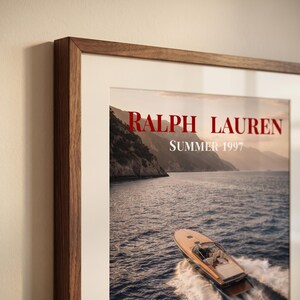 May include: A framed wall art print featuring a wooden speedboat moving through dark blue water near a rocky coastline. The text RALPH LAUREN and SUMMER 1997 are printed in red and white at the top. The frame is made of dark brown wood.