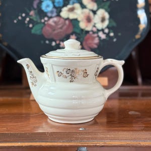 May include: Cream-colored ceramic teapot with a lid, featuring gold floral accents. The teapot has a curved spout and handle. It sits on a wooden surface, with a decorative tray in the background.