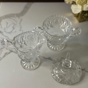 May include: Clear glass creamer, sugar bowl, and lid set. The creamer has a handle and spout, while the sugar bowl has two handles. The lid features a pointed handle. All pieces have a cut glass design.