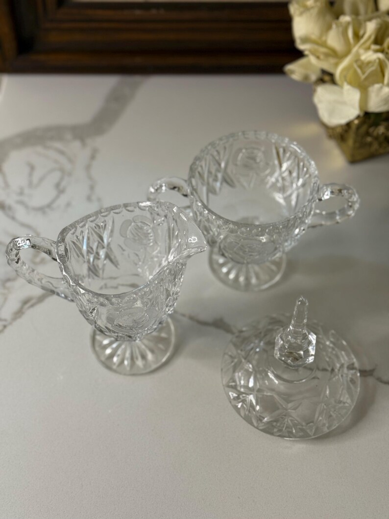 May include: Clear glass creamer, sugar bowl, and lid set. The creamer has a handle and spout, while the sugar bowl has two handles. The lid features a pointed handle. All pieces have a cut glass design.