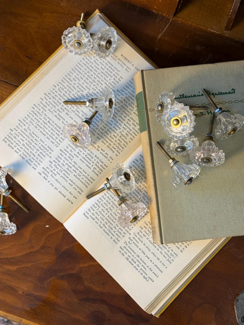 May include: Clear, faceted glass cabinet knobs with brass hardware are arranged on open books. The knobs have a vintage, antique aesthetic. The books are open to reveal printed text.