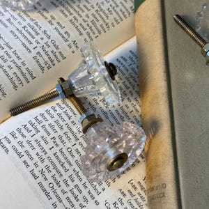 May include: Clear glass cabinet knobs with brass hardware are displayed on open books. The knobs have a faceted design, reflecting light. The background includes book pages and a book spine, creating a vintage aesthetic.