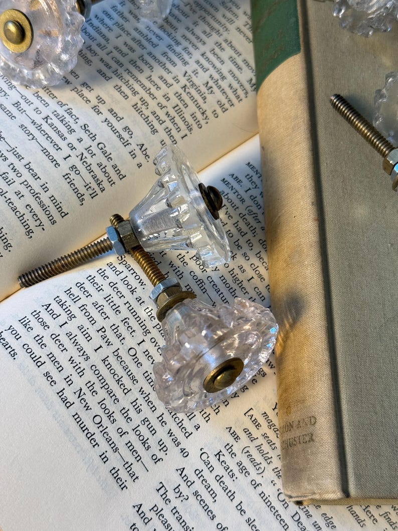 May include: Clear glass cabinet knobs with brass hardware are displayed on open books. The knobs have a faceted design, reflecting light. The background includes book pages and a book spine, creating a vintage aesthetic.