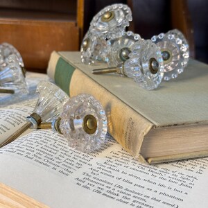 May include: Clear glass cabinet knobs with a faceted design and brass hardware are arranged on an open book. The knobs have a vintage aesthetic, with a classic, decorative style. The book has aged pages and a green spine.