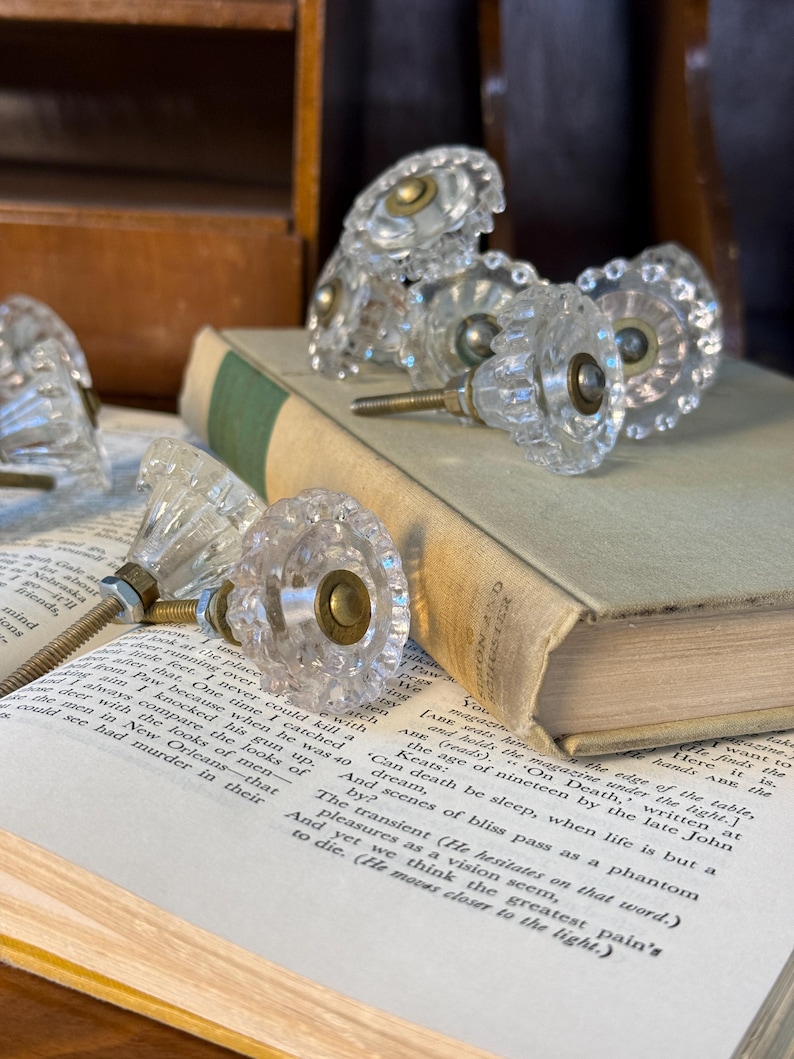May include: Clear glass cabinet knobs with a faceted design and brass hardware are arranged on an open book. The knobs have a vintage aesthetic, with a classic, decorative style. The book has aged pages and a green spine.
