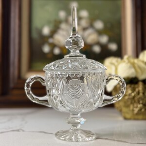 May include: A clear, vintage glass sugar bowl with a lid and two handles. The bowl features a rose design and diamond-cut detailing. The sugar bowl is on a pedestal base and is likely used for serving sugar or other condiments.