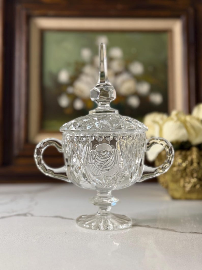 May include: A clear, vintage glass sugar bowl with a lid and two handles. The bowl features a rose design and diamond-cut detailing. The sugar bowl is on a pedestal base and is likely used for serving sugar or other condiments.