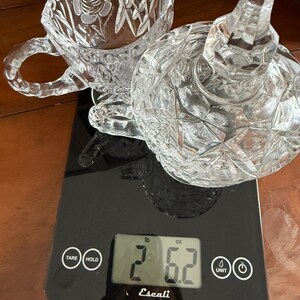 May include: A clear, cut-glass teacup and a lid with a stem are on a black digital scale. The teacup has a rose design. The scale reads 2 pounds and 6.2 ounces. The brand name "Escali" is visible on the scale.