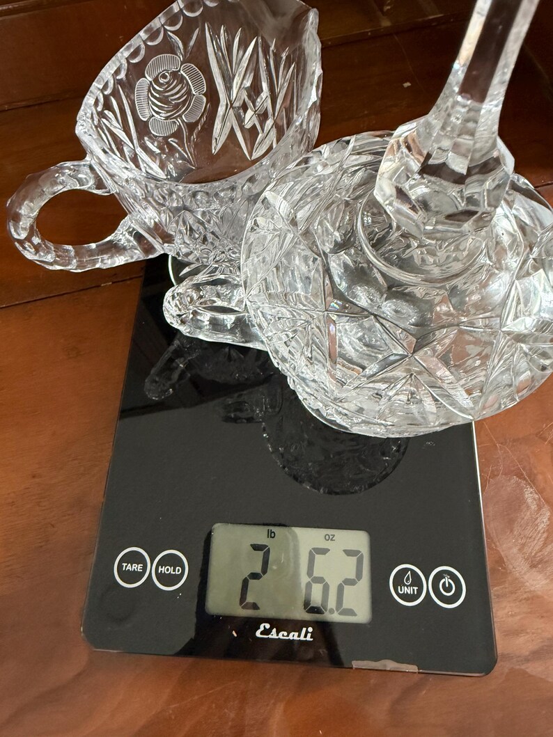 May include: A clear, cut-glass teacup and a lid with a stem are on a black digital scale. The teacup has a rose design. The scale reads 2 pounds and 6.2 ounces. The brand name "Escali" is visible on the scale.
