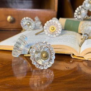 May include: Clear glass cabinet knobs with a scalloped edge and brass hardware. The knobs are arranged on a wooden surface with an open book in the background. The knobs have a vintage, antique aesthetic.