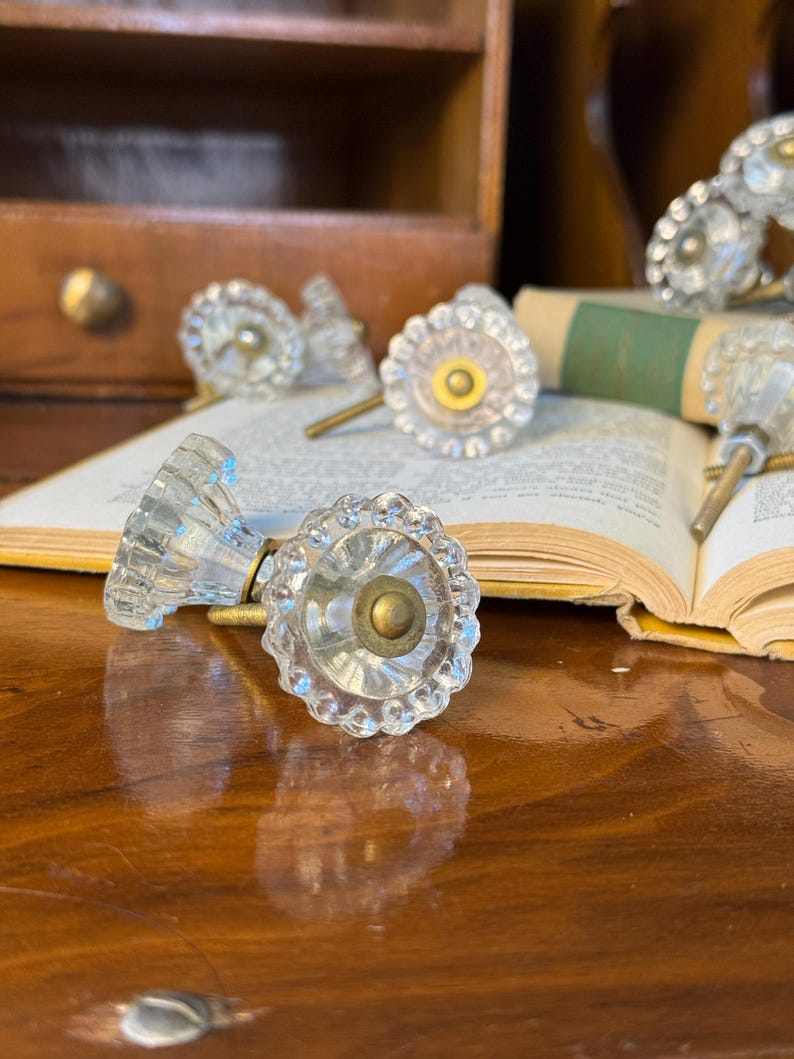 May include: Clear glass cabinet knobs with a scalloped edge and brass hardware. The knobs are arranged on a wooden surface with an open book in the background. The knobs have a vintage, antique aesthetic.