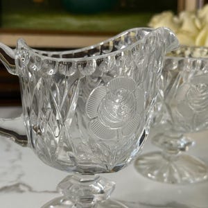 May include: Clear glass creamer with a rose design. The creamer has a handle and a spout. The base is a pedestal design. The creamer is part of a set. The glass has a diamond pattern.